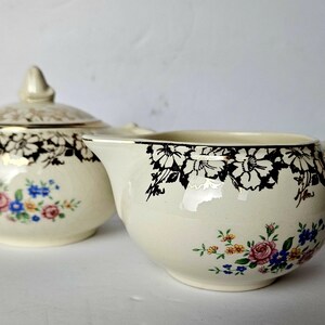 Peut inclure: Sucrier en céramique crème avec couvercle et crémier. Les deux pièces présentent des motifs floraux noirs sur les bords et des bouquets floraux colorés. Le sucrier a un couvercle et des poignées dorés.