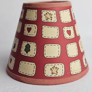 May include: A lampshade with a red background, decorated with beige rectangles. Each rectangle features a heart, star, or tree illustration. The top and bottom edges are trimmed with a matching red fabric. A decorative item.