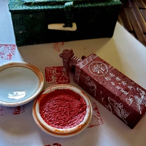 Puede incluir: Un juego de sellos de tinta roja vintage con una caja decorativa. El juego incluye una almohadilla de tinta roja, un tintero de cerámica y un sello de piedra roja con caracteres grabados. La caja es verde con un cierre.