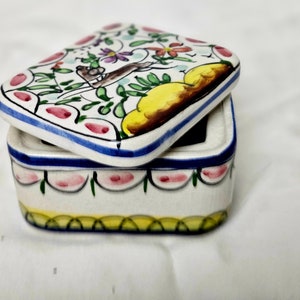 Könnte beinhalten: Rechteckige Schmuckschatulle aus Keramik mit Deckel. Die Schachtel und der Deckel sind weiß mit handgemalten Blumen- und Blattmotiven in Rosa, Grün, Gelb und Braun. Der Deckel hat einen blauen Rand.