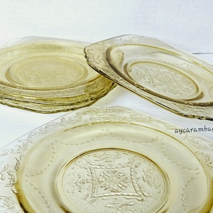 May include: A stack of six yellow-tinted glass plates with a square shape and ornate, embossed floral designs. The plates have a scalloped edge and a central embossed pattern. The plates are arranged on a white surface.