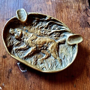 May include: A brass ashtray with an embossed design of a dog running through tall grass. The ashtray is oval shaped and has a raised edge.