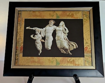 May include: Framed artwork depicting a classical relief sculpture of three figures. The figures are white against a black background, framed by a gold and map-like design, and encased in a black frame. The artwork is displayed on a black easel.