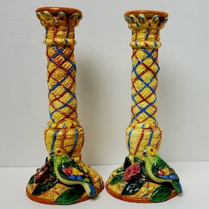 Puede incluir: Un par de candelabros de cerámica con un patrón tejido amarillo, rojo y azul. Cada candelabro presenta un loro con plumas verdes y azules y una flor roja.
