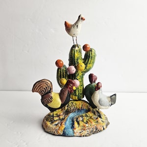 May include: A colorful, hand-painted cast iron figurine featuring a cactus with a chicken perched on top. Two roosters and a hen are at the base. The base is painted to resemble a desert landscape with a blue river.
