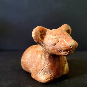May include: A small, brown, clay sculpture of a resting animal, possibly a rodent, with large ears and a detailed face. The sculpture has a rough texture and is set against a dark background.