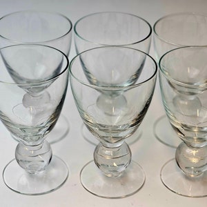 Może przedstawiać: Sześć przezroczystych kieliszków do wina. Każdy kieliszek ma zaokrągloną czaszę, dekoracyjną kulistą podstawę i okrągłą stopkę. Kieliszki są ułożone w dwóch rzędach po trzy, podkreślając ich elegancki design i przejrzystość.