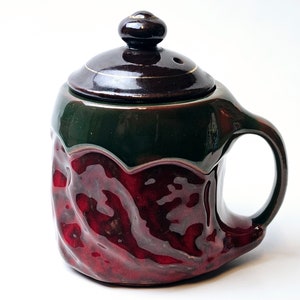 Puede incluir: Una taza de cerámica con una tapa marrón oscura y un esmalte verde y rojo. La taza tiene una superficie texturizada, irregular y un asa.