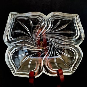 May include: Clear glass serving dish with a four-sectioned design. Each section features a swirling, stylized floral pattern. The dish is displayed on a dark surface, showcasing its intricate details and reflective qualities.