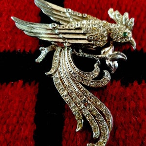 Puede incluir: Broche plateado con forma de pájaro, adornado con brillantes transparentes y un ojo verde esmeralda. El pájaro tiene las alas extendidas y una cola larga y fluida. El broche está sobre un fondo a cuadros rojo y negro.