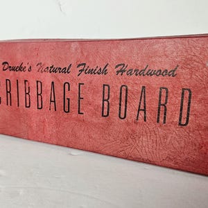 May include: Red cribbage board box with the text "Druke's Natural Finish Hardwood CRIBAGE BOARD". The box has a textured surface and a knight chess piece logo. A classic game for family fun.