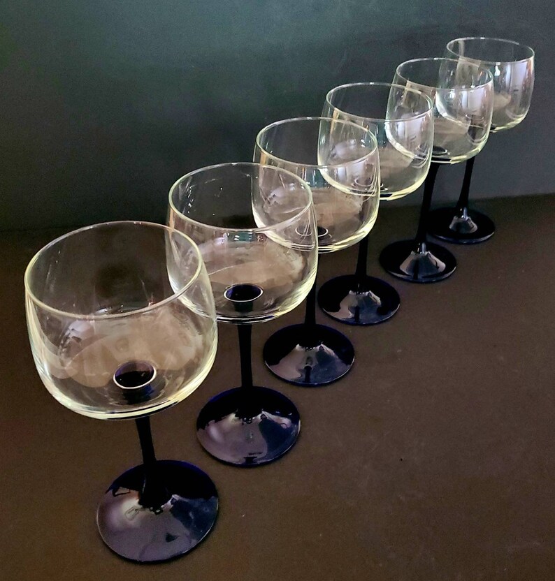 May include: Set of six clear glass wine glasses with dark blue stems and bases. The glasses are arranged in a row, with the stems pointing to the right.