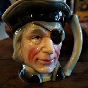 May include: A ceramic mug shaped like a pirate's head, featuring a black eye patch, a tricorn hat, and a handle. The mug has a painted face with rosy cheeks and a stern expression. The mug is a decorative item.