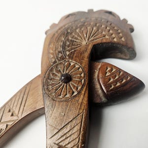 May include: Close-up of a vintage wooden nutcracker with intricate carved details. The nutcracker features a dark brown finish and a lever-style design. The head and handle are adorned with floral and geometric patterns.