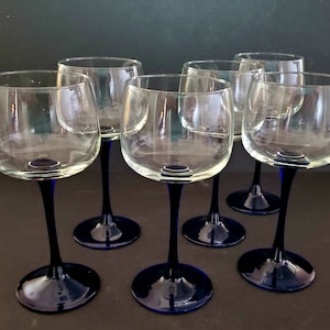 May include: Set of six clear glass wine glasses with blue stems. The glasses are arranged in a semi-circle on a dark surface.