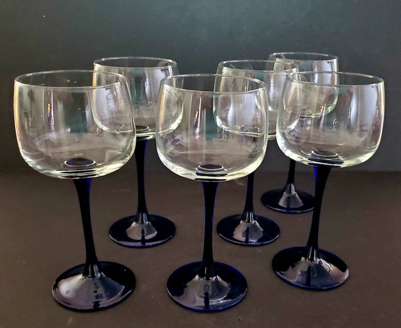 May include: Set of six clear glass wine glasses with blue stems. The glasses are arranged in a semi-circle on a dark surface.