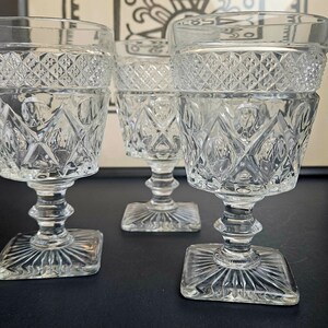 May include: Three clear, vintage crystal goblets with diamond and geometric patterns. Each goblet has a square base, a detailed stem, and a decorative rim. The glasses are arranged on a dark surface, with a framed artwork in the background.