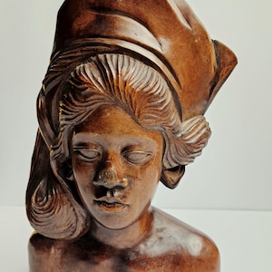 Peut inclure: Buste en bois sculpté d'une femme aux yeux fermés, portant une coiffe. La sculpture est de couleur brun chaud, avec une sculpture détaillée des cheveux et des traits du visage. Le buste est posé sur un fond blanc.