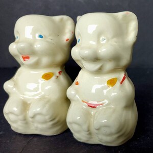 May include: A pair of white ceramic salt and pepper shakers shaped like bears. The bears have blue eyes, red mouths, and yellow and red painted details.