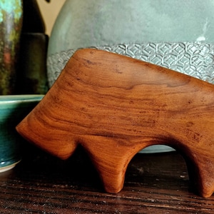 Könnte beinhalten: Handgefertigte Holzskulptur eines Bisons mit einem satten, rotbraunen Finish. Der Bison ist mit glatten Linien geschnitzt und hat eine markante Form. Die Skulptur wird auf einer dunklen Holzoberfläche mit Keramikvasen im Hintergrund präsentiert.
