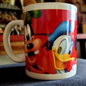 May include: A red ceramic mug with a cartoon image of Mickey Mouse and Donald Duck. The mug has a white handle and a white rim.