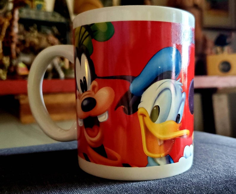 May include: A red ceramic mug with a cartoon image of Mickey Mouse and Donald Duck. The mug has a white handle and a white rim.