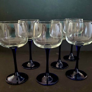 May include: Six clear glass wine glasses with blue stems and bases. The glasses are arranged in a semi-circle on a dark surface.