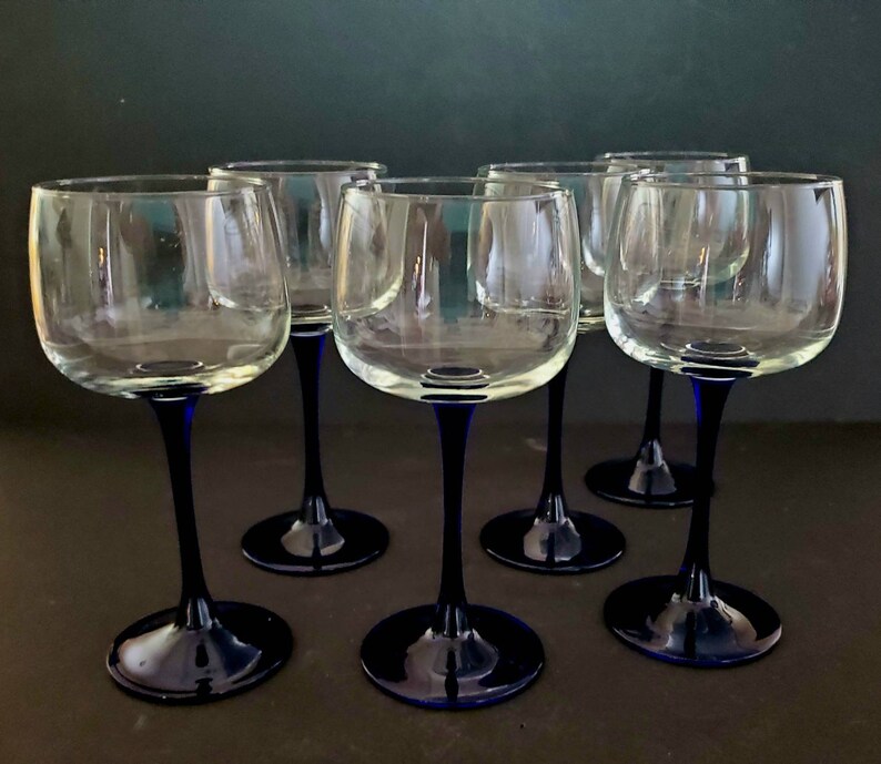 May include: Six clear glass wine glasses with blue stems and bases. The glasses are arranged in a semi-circle on a dark surface.