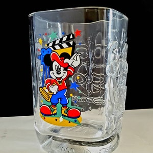 May include: A clear glass cup with a Mickey Mouse design. Mickey is wearing a red hat and blue pants and is holding a trumpet. The cup has a 2000 Walt Disney World logo on the bottom.