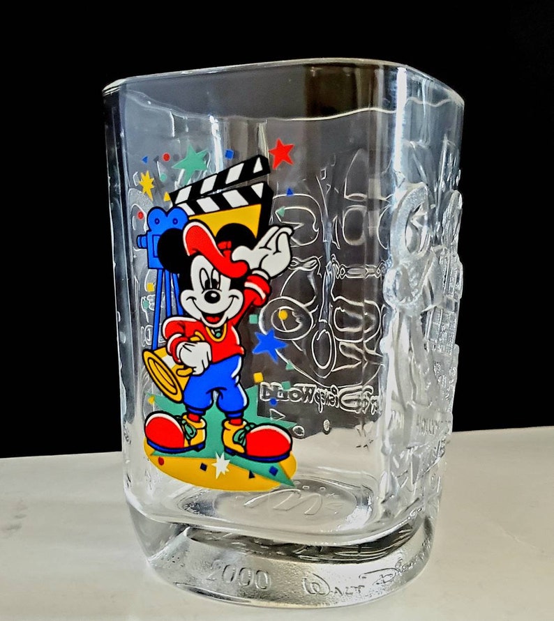 May include: A clear glass cup with a Mickey Mouse design. Mickey is wearing a red hat and blue pants and is holding a trumpet. The cup has a 2000 Walt Disney World logo on the bottom.