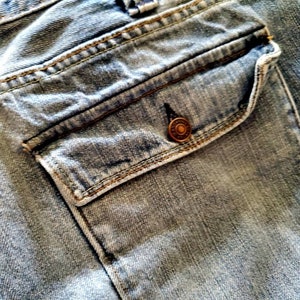 May include: Close-up of a blue denim pocket with a metal button. The pocket is sewn with a contrasting thread.