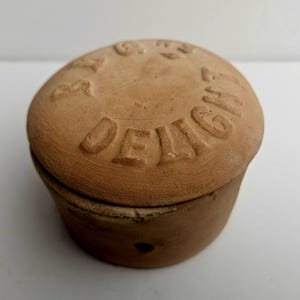 Può includere: Un piccolo contenitore rotondo color terracotta con coperchio. Il coperchio è impresso con la parola "DELIGHT" in maiuscolo. Il contenitore ha un design semplice e rustico, adatto per riporre piccoli oggetti.