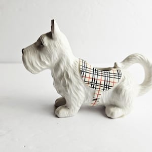 Könnte beinhalten: Eine weiße Keramikfigur eines Scottish Terrier-Hundes. Der Hund hat ein strukturiertes Fell und einen kleinen Sattel mit einem schwarz-rot-weiß karierten Muster. Die Figur sitzt auf ihren vier Beinen und hat einen gebogenen Schwanz.