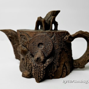 Puede incluir: Una tetera de madera marrón detallada, tallada para parecerse a un tocón de árbol. La tetera presenta un asa, un pico y una tapa con un elemento decorativo. Un diseño similar a una moneda está tallado en la parte delantera. El diseño general es rústico y artístico.