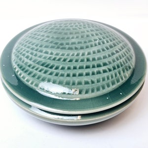 Puede incluir: Un plato de cerámica de color verde azulado con tapa y un patrón geométrico texturizado en la parte superior. El plato es redondo y tiene una tapa ligeramente abovedada.
