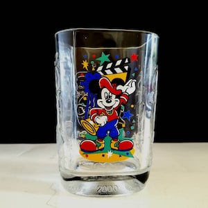 May include: A clear glass cup with a Mickey Mouse design. Mickey is wearing a red hat and holding a microphone. The cup has a blue, yellow, and green design with stars and confetti. The bottom of the cup says "Disney" and "2000".