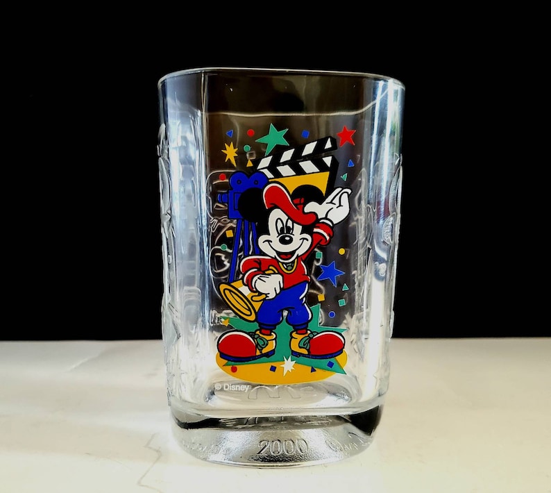 May include: A clear glass cup with a Mickey Mouse design. Mickey is wearing a red hat and holding a microphone. The cup has a blue, yellow, and green design with stars and confetti. The bottom of the cup says "Disney" and "2000".