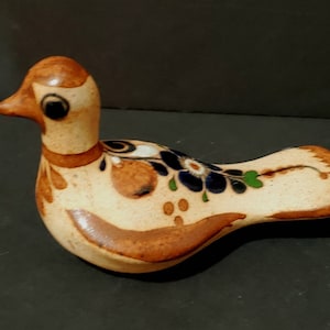 Puede incluir: Figura de pájaro de cerámica con cuerpo beige, detalles marrones y diseños florales en azul, blanco y verde. El pájaro tiene un cuerpo largo y curvado y una cabeza pequeña con ojos negros y pico marrón.
