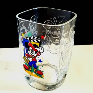 May include: A clear glass cup with a Mickey Mouse design. Mickey is wearing a red hat and holding a clapperboard. The cup has the words "Hollywood" and "Disney" on it.