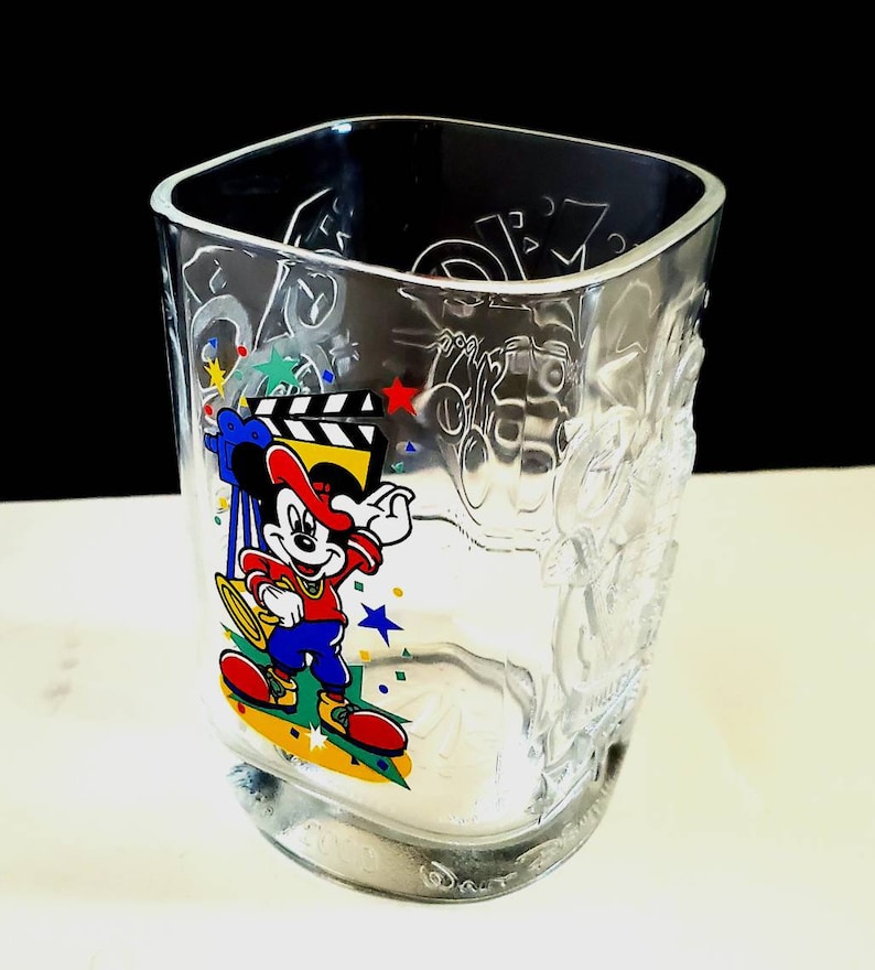 May include: A clear glass cup with a Mickey Mouse design. Mickey is wearing a red hat and holding a clapperboard. The cup has the words "Hollywood" and "Disney" on it.