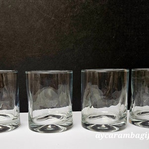 May include: Four clear glass tumblers on a white surface. The tumblers are empty and have a rounded bottom.