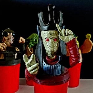 以下が含まれることがあります： 赤い目をした緑と灰色のエイリアンのフィギュアが、黒と赤のローブと王冠を身に着けています。フィギュアは右手を顔の前に上げています。フィギュアは赤いカップの上に立っています。