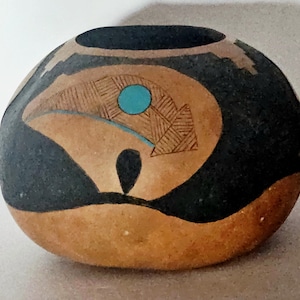 May include: A round, hand-painted ceramic pot with a black exterior and a tan-coloured base. The pot features a stylized bear design with a turquoise circle and geometric patterns. The opening is black.