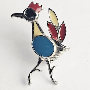 May include: A vintage-style brooch in the shape of a bird. The brooch features a silver-tone metal body with a red comb, black eye, yellow and red tail feathers, and a blue oval body. The bird is standing with its legs slightly bent.