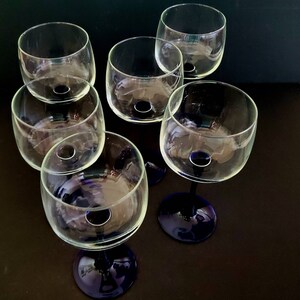 May include: Six clear glass wine glasses with blue stems. The glasses are arranged in a circle on a dark surface.