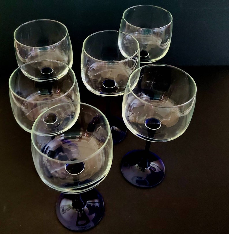 May include: Six clear glass wine glasses with blue stems. The glasses are arranged in a circle on a dark surface.