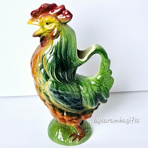 May include: A green, yellow, and red ceramic rooster pitcher. The rooster is standing on a green base and has a spout on the side of its head.