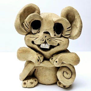 May include: A ceramic figurine of a smiling mouse with large eyes and a toothy grin. The mouse is sitting with its arms crossed and its tail curled around its body. The figurine is painted in shades of brown and beige.