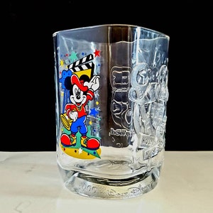 May include: A clear glass cup with a Mickey Mouse design. The cup features Mickey Mouse in a red shirt and blue pants, holding a trumpet. The cup also has a film reel and stars on it. The text "Hollywood Studios" is on the cup.
