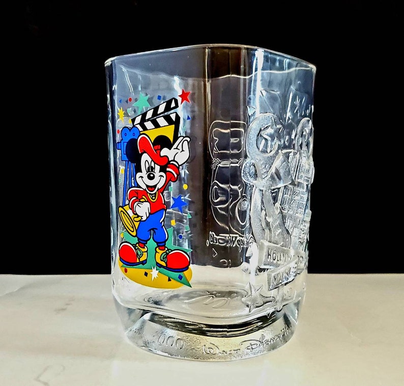 May include: A clear glass cup with a Mickey Mouse design. The cup features Mickey Mouse in a red shirt and blue pants, holding a trumpet. The cup also has a film reel and stars on it. The text "Hollywood Studios" is on the cup.